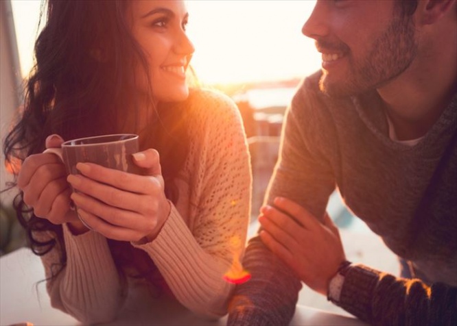 Couple drinking coffee and talking. They are looking at each other at sunrise or sunset. He has a beard.