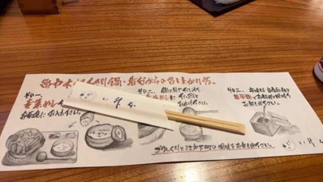 Japanese menu sheets with drawings of dishes and handwritten text on a wooden table, with chopsticks resting on top.