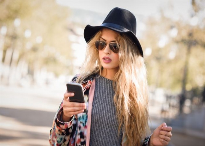 Young woman looking at her smartphone in urban background. Girl wearing jacket, hat, sweater and sunglasses.