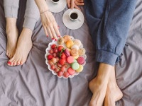 Young caucasian couple having romantic breakfast in bed. Closeup of female and male feet, two cups of coffee, fruits and colorful biscuits.