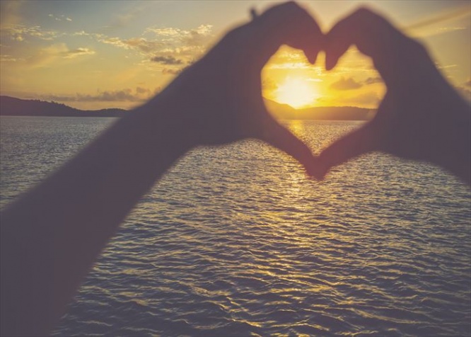 Woman making a heart shape with her hands. It is sunset behind her hands with light reflecting off the ocean. Focus is on the background