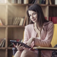 Beautiful young smart woman sits with the electronic book in the library on a chair
