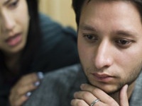 Indoor image of sad young man consoled by a young woman. Selective focus is on young man and young woman is defocused in the background, she is holding his shoulders from behind and supporting him. Two people, horizontal composition with selective focus.