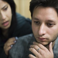 Indoor image of sad young man consoled by a young woman. Selective focus is on young man and young woman is defocused in the background, she is holding his shoulders from behind and supporting him. Two people, horizontal composition with selective focus.