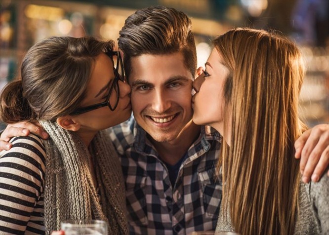 Three young people enjoying in a cafe.  Young women are kissing in cheek a man who is embracing them and looking at camera.