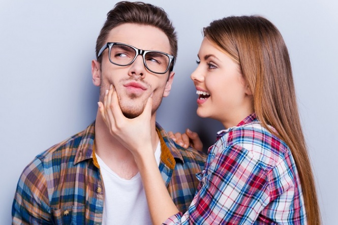 Handsome young man grimacing while his girlfriend touching his face while standing against grey background