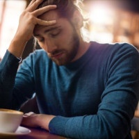 Young man with eyes closed having a headache while sitting in a cafe and holding his head in pain.