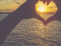 Woman making a heart shape with her hands. It is sunset behind her hands with light reflecting off the ocean. Focus is on the background