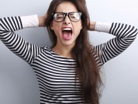 Anger young business woman in glasses strong screaming with wild open mouth and holding head the hands on blue background
