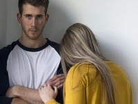 Young woman crying on her partners shoulder. He is looking at the camera with his arms folded.