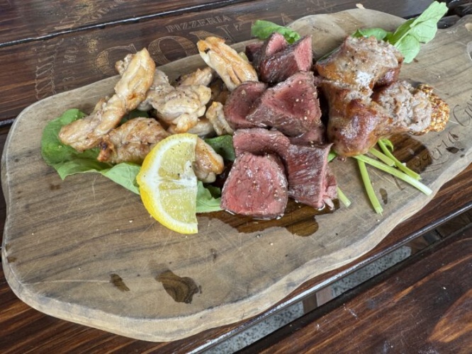 Assorted grilled meats (steak and chicken) on a wooden board with greens and a lemon wedge.