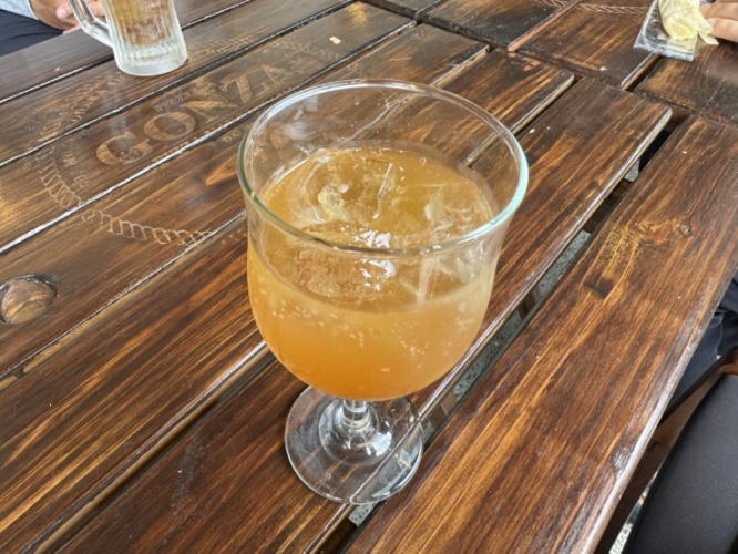 Tall stemmed glass of orange iced drink with ice on a rustic wooden table; hands and napkins visible at the edges.